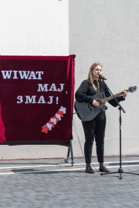 Dziewczyna trzymająca gitarę (śpiewająca?) na tle napisu "Wiwat maj, 3 maj"