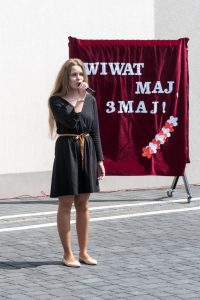 Dziewczyna trzymająca mikrofon (śpiewająca?) na tle napisu "Wiwat maj, 3 maj"